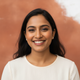 Smiling South Asian woman in her late 20s, a natural portrait headshot, against a soft background of terracotta and white.